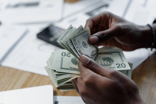 courses-02-free-img A close-up of hands counting US dollar bills on a wooden table with documents and a smartphone nearby.